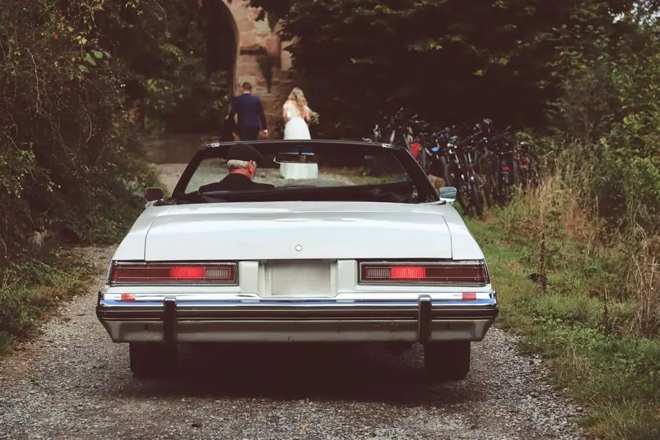 A vintage car parked on a gravel path, with a bride and groom walking ahead.