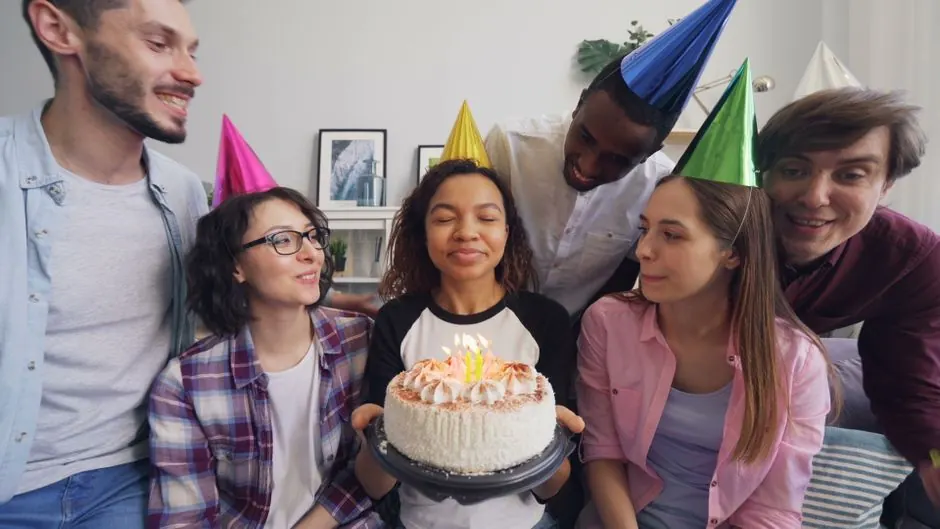 Group of friends celebrating a birthday with a cake and party hats indoors.